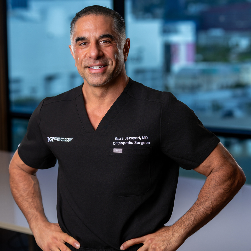 A smiling man in black medical scrubs stands with his hands on his hips in an office setting. His shirt reads Reza Jazayeri, MD Orthopedic Surgeon. Large windows and a blurred cityscape are visible in the background.