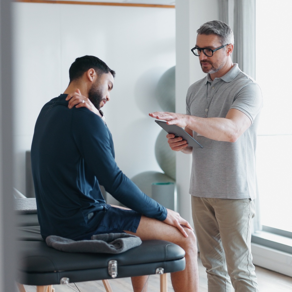 Physiotherapy, tablet and shoulder pain with people in clinic for rehabilitation, healthcare or consulting. Support, medical and chiropractor with man and athlete for healing, advice or report.
