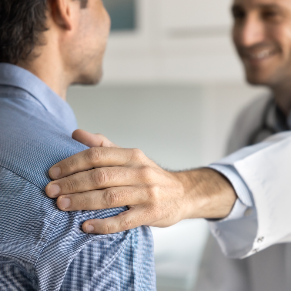 Smiling doctor in uniform speaking with male patient, touches his shoulder as gesture of encouragement, care and compassion, share good news about disease recovery, provide emotional support, close up