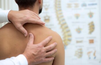 A healthcare professional examines a mans bare shoulder and upper back with a spine diagram blurred in the background.