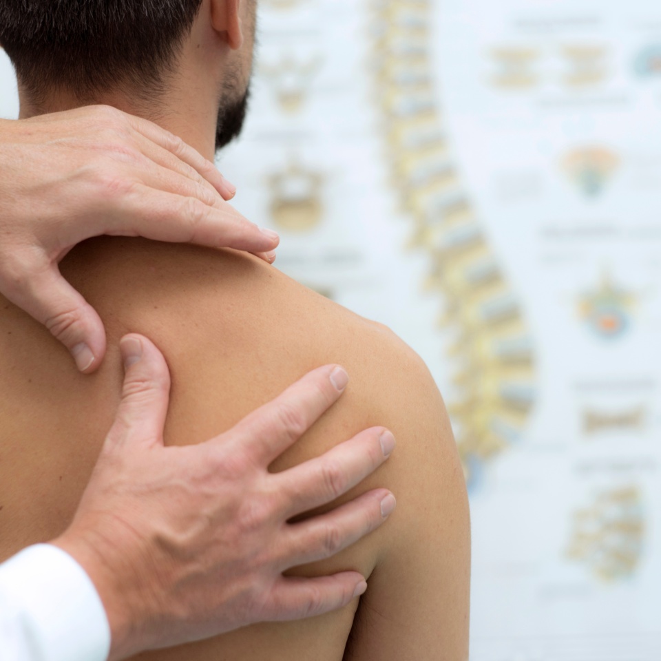 A healthcare professional examines a mans bare shoulder and upper back, with a blurred diagram of the human spine visible in the background.
