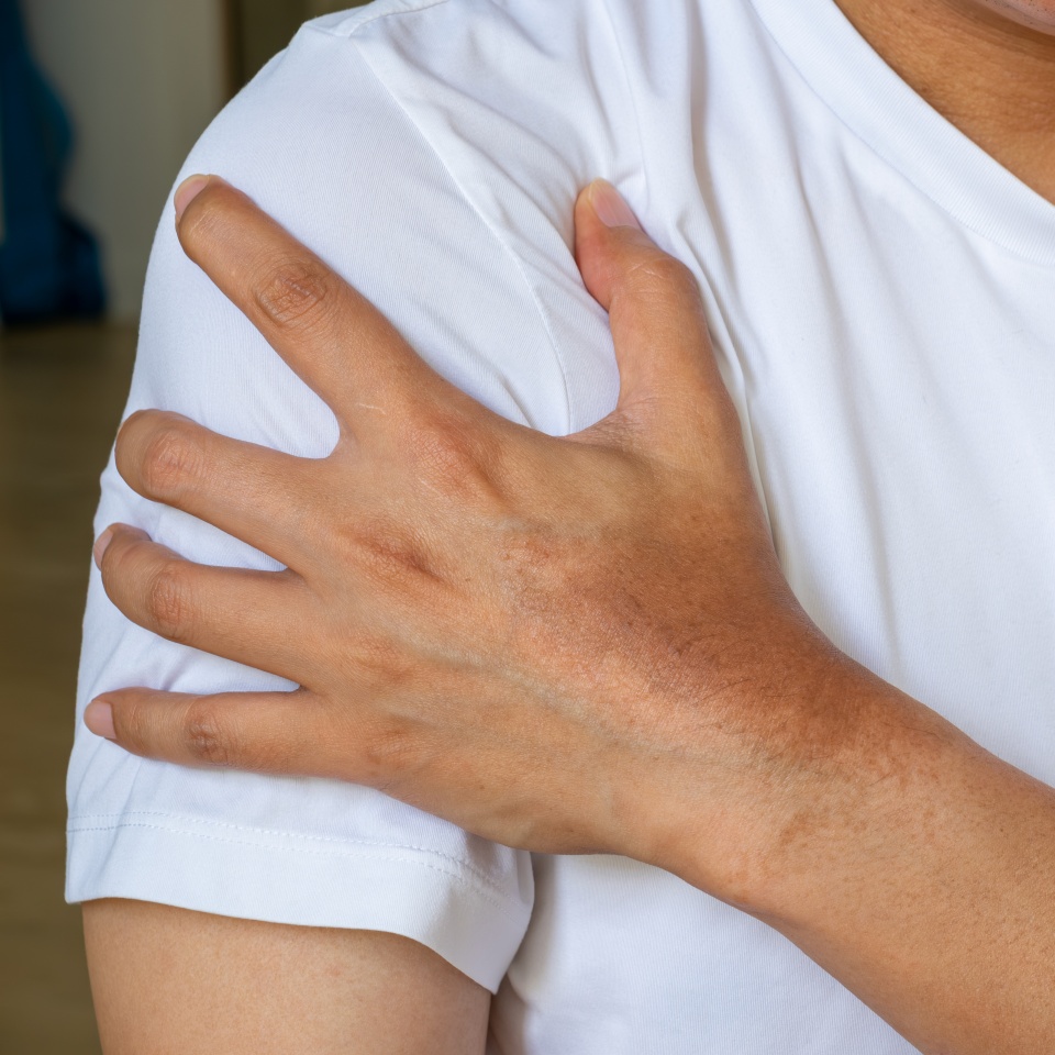 A person wearing a white shirt holds their left shoulder with their right hand, suggesting shoulder pain or discomfort.