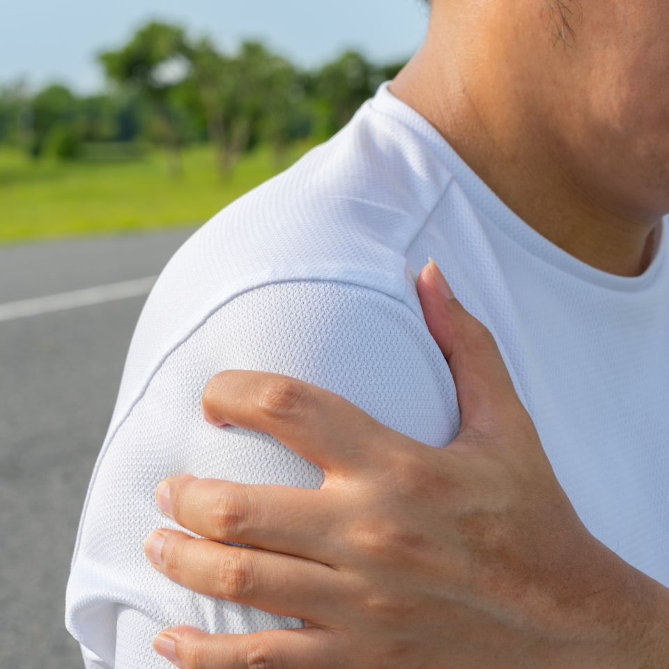A person wearing a white shirt is holding their left shoulder with their right hand, appearing to be in pain or discomfort, with an outdoor background of grass and trees.
