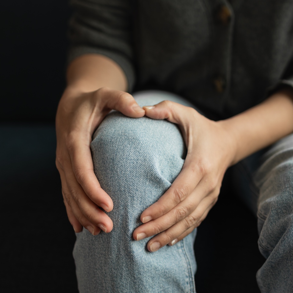 A person wearing light blue jeans holds their knee with both hands, suggesting discomfort or pain. The background is dark, and the persons face is not visible.