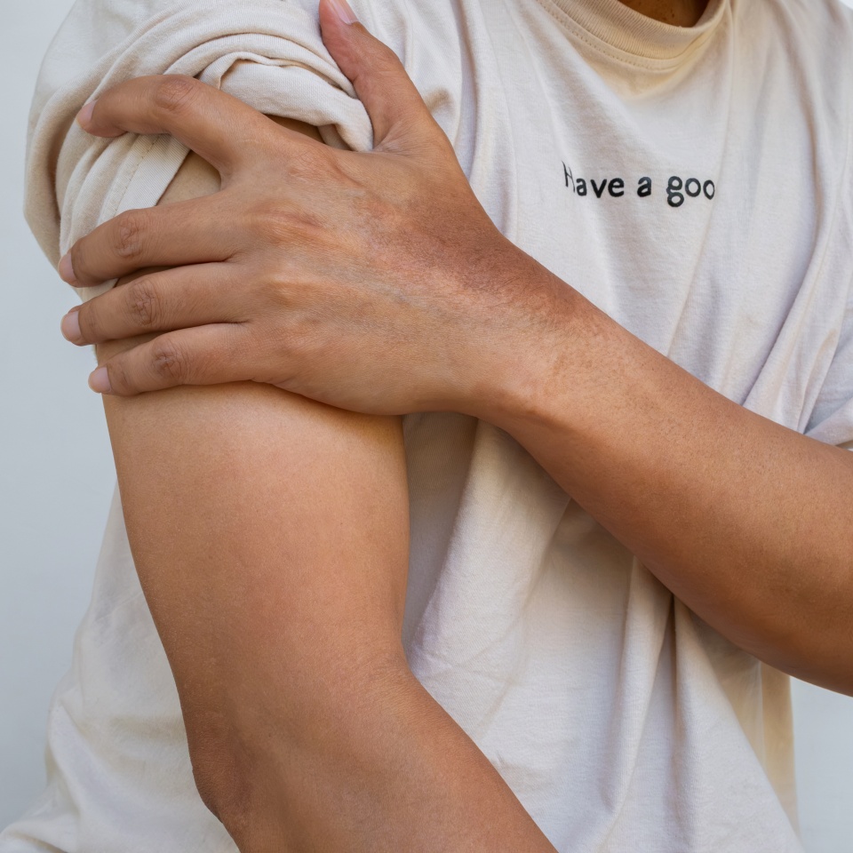 A person wearing a light-colored t-shirt holds their upper arm with their opposite hand, as if feeling pain or soreness. The t-shirt has partially visible text that reads have a goo.