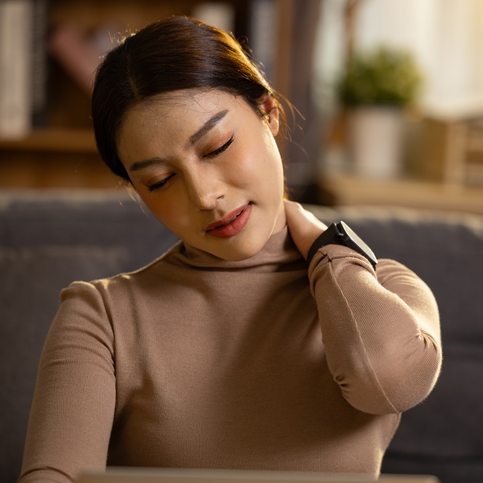 Exhausted young Asian woman suffering from neck pain and shoulder stiffness touches sore muscle while working late feeling office syndrome stress at home living room on cozy sofa looking unhappy
