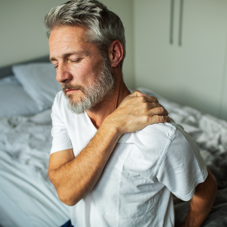 A middle-aged man with gray hair and a beard sits on a bed, wearing a white shirt, with his eyes closed and his hand on his shoulder, appearing to be in pain or discomfort.