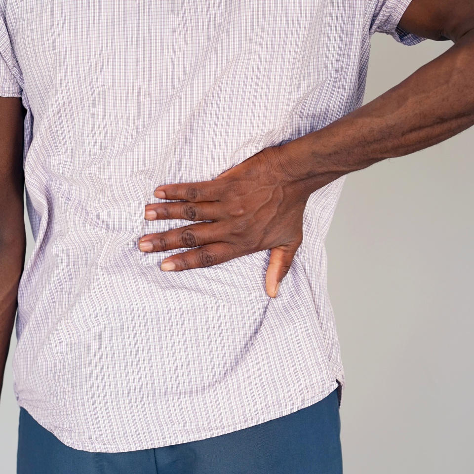 Close-up view of a young man with pain in kidneys on gray background. Young man with back ache clasping her hand to her lower back. Man suffering from ribbing pain, waist pain.
