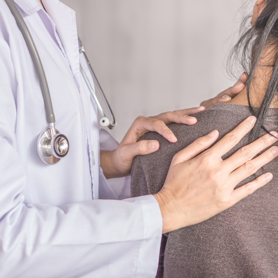 A doctor in a white coat with a stethoscope gently places hands on a patients shoulder, offering comfort and support. The patient is seen from behind, wearing a brown sweater.