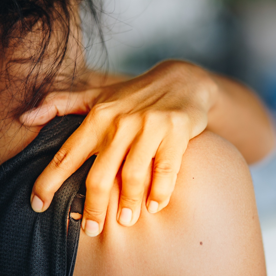 A person with dark hair touches their left shoulder, appearing to be in discomfort or pain, with their hand gripping the shoulder of a sleeveless shirt. The background is softly blurred.