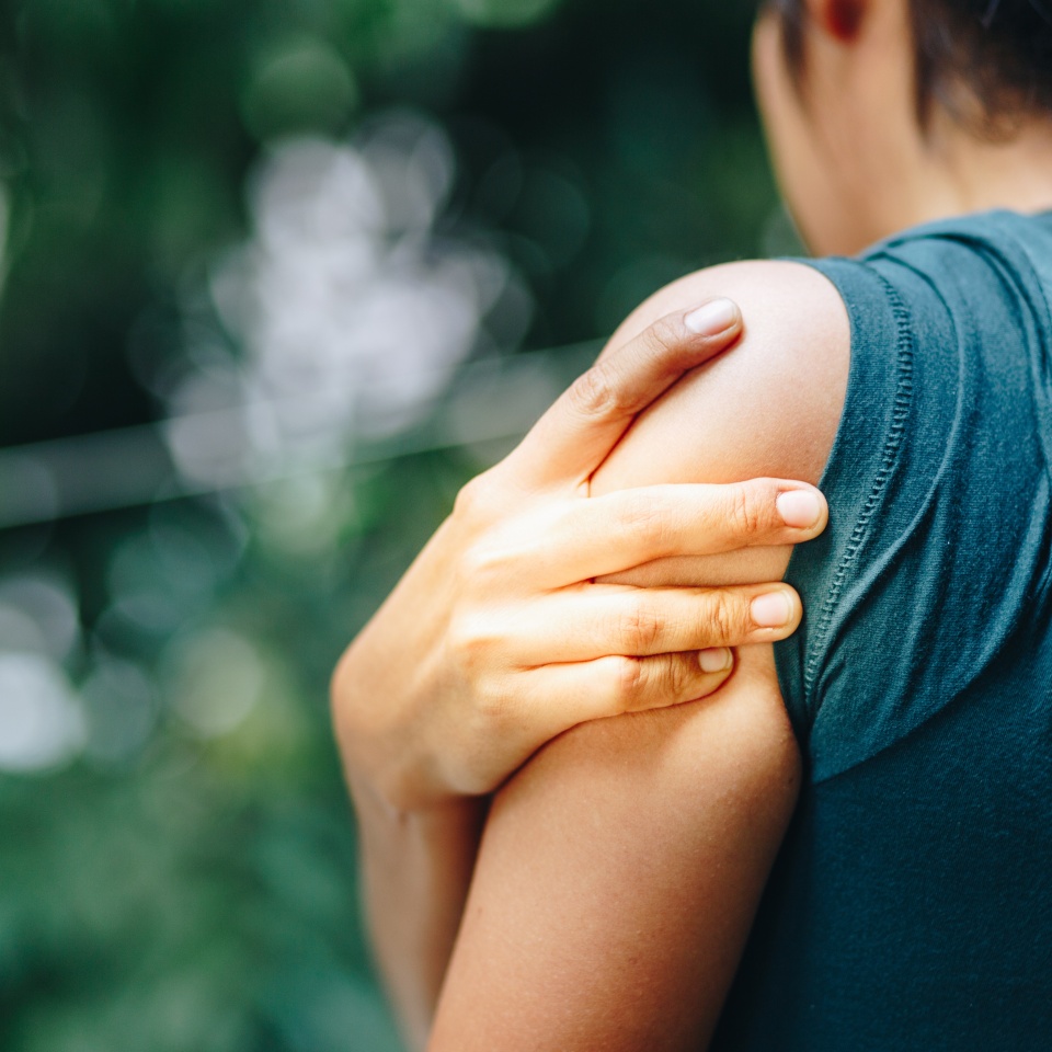 A person wearing a dark shirt is seen from behind, holding their left shoulder with their right hand, as if experiencing pain or discomfort. The background is blurred with green foliage.