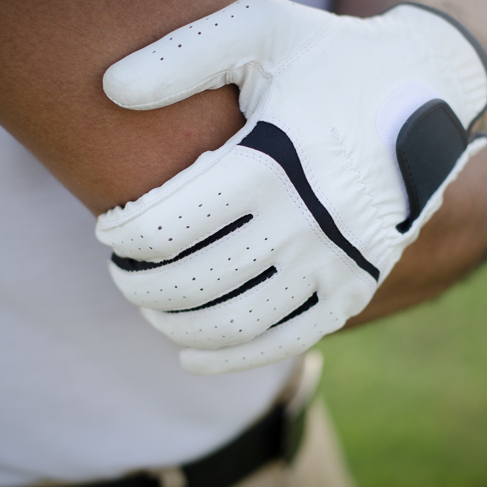 A person wearing a white sports glove holds their elbow with their other hand, possibly indicating pain or discomfort, while standing outdoors.