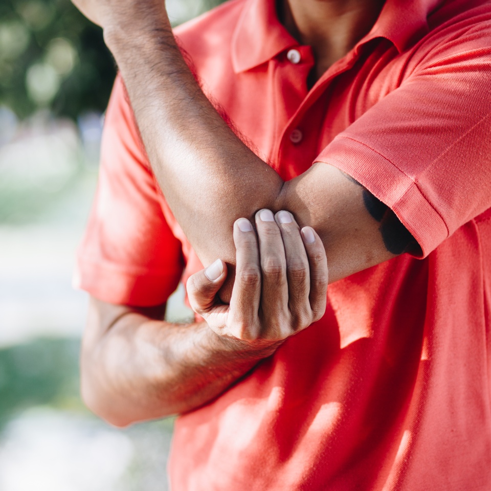 A person in a red shirt holds their left elbow with their right hand, appearing to be in discomfort or pain, outdoors with blurred greenery in the background.