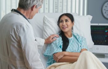 Supportive and caring older doctor with hand on shoulder of recovering female patient in hospital bed. Japanese woman getting good news from senior Caucasian physician