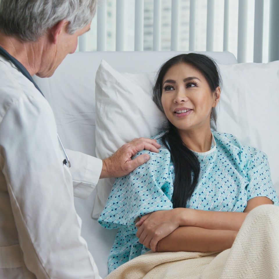 Supportive and caring older doctor with hand on shoulder of recovering female patient in hospital bed. Japanese woman getting good news from senior Caucasian physician