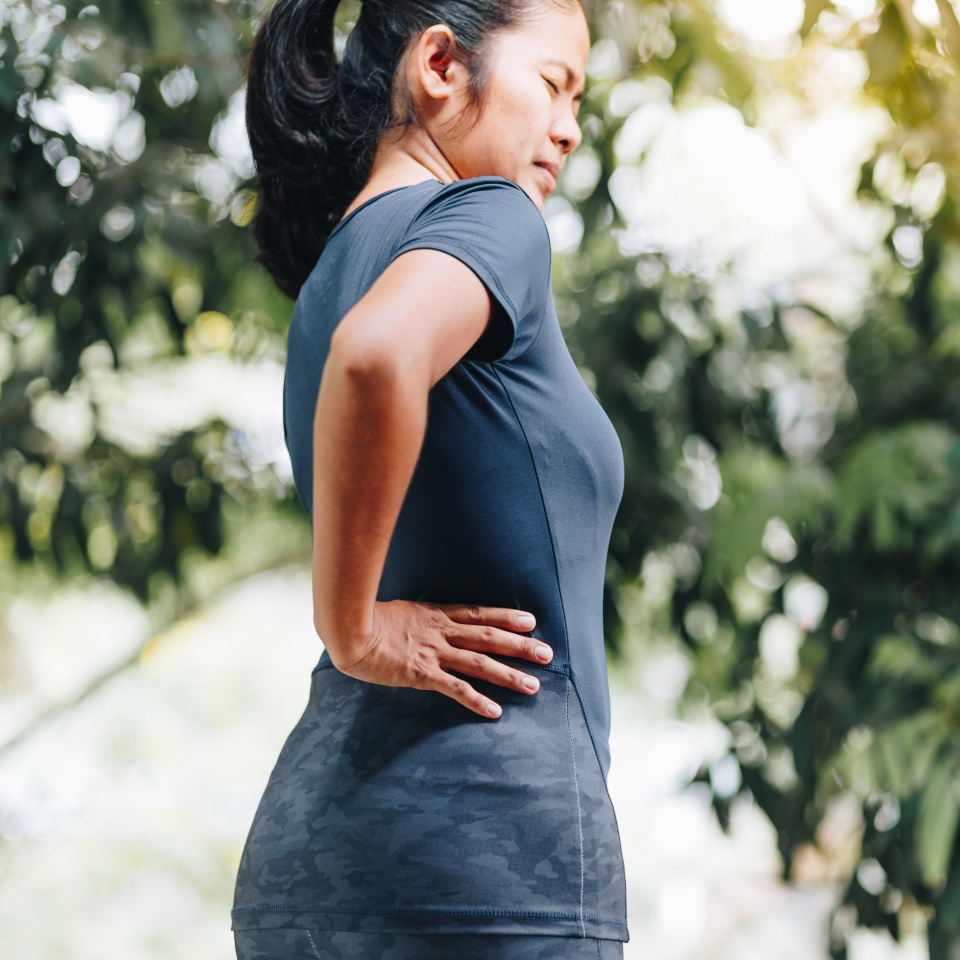 A woman wearing athletic clothing stands outdoors, holding her lower back with one hand and appearing to be in pain, possibly experiencing back discomfort. Lush green foliage is visible in the background.