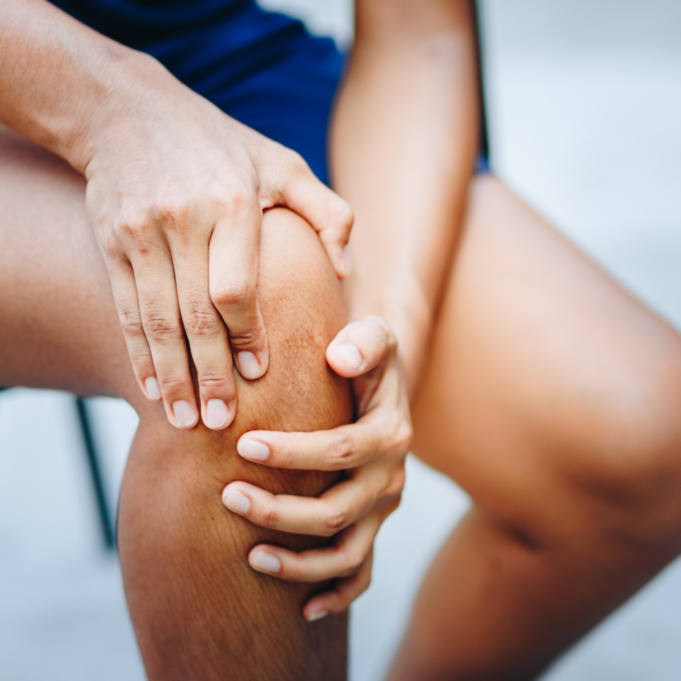 A person sitting and holding their knee with both hands, appearing to experience knee pain or discomfort.