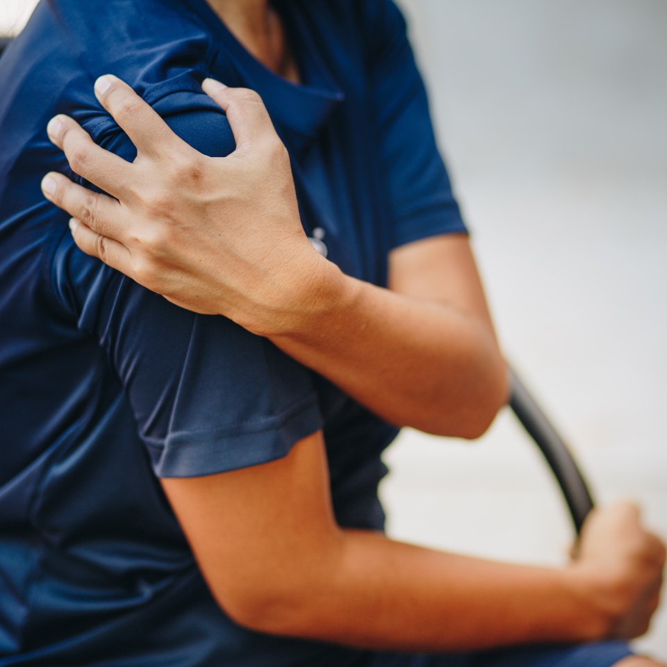 A person wearing a navy blue shirt sits and holds their left shoulder with their right hand, appearing to be in pain or discomfort.