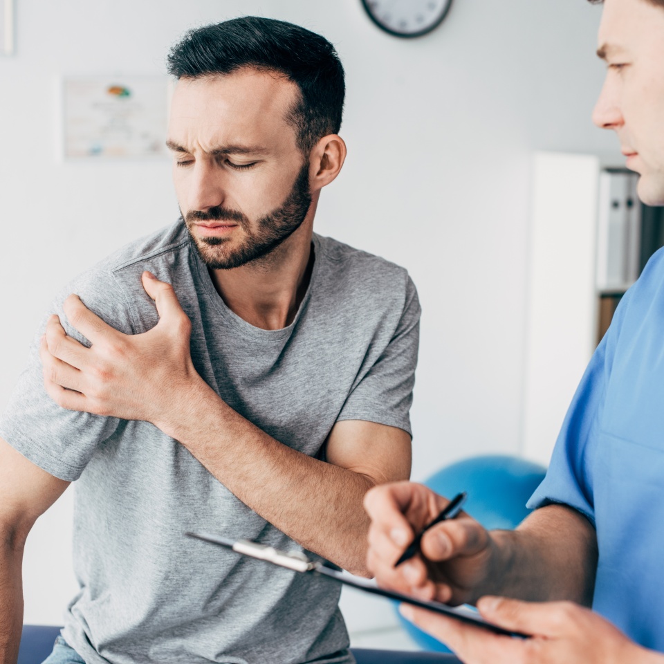 Selective focus of patient touching his shoulder and suffering from pain and doctor writing prescription in massage cabinet at clinic