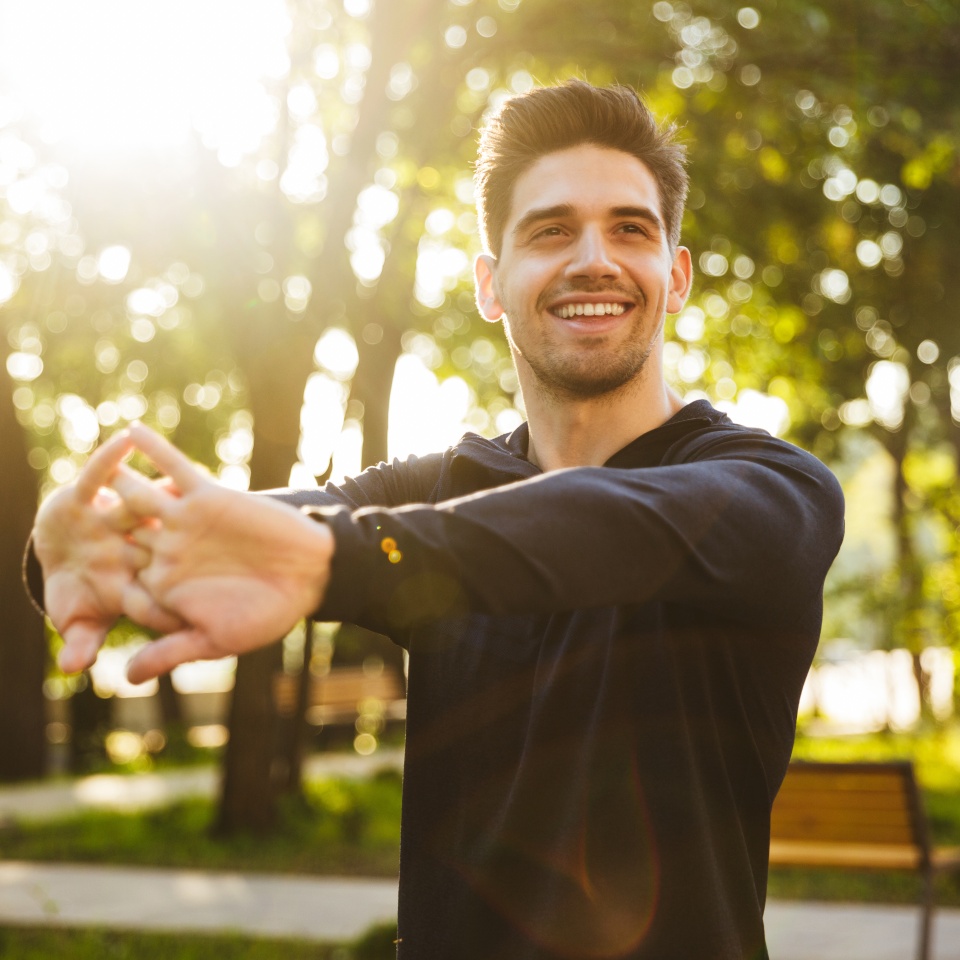 Image of a handsome smiling young sports fitness man standing in green park nature make stretching exercises for arms.