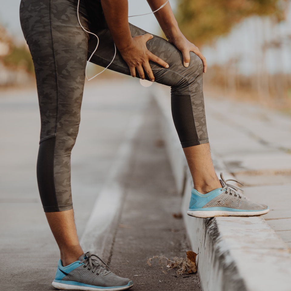 A person in athletic wear stands on a curb, holding their knee as if in pain or discomfort, with one leg bent and the other resting on the curb. The background shows an empty street lined with trees.