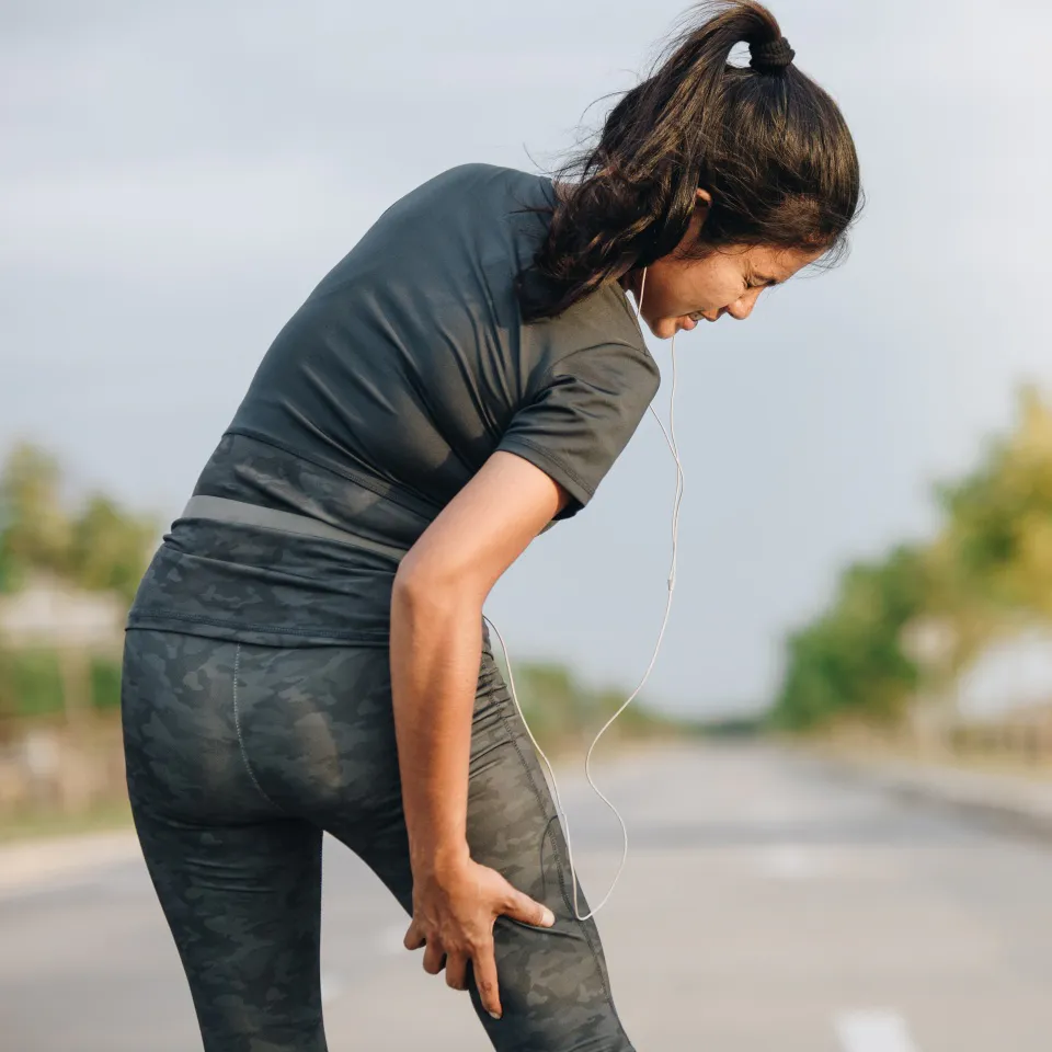 A woman in athletic wear stands on a road, holding her thigh and appearing to be in pain, possibly from a muscle cramp or injury. She has earbuds in and is outdoors, with trees lining the road.