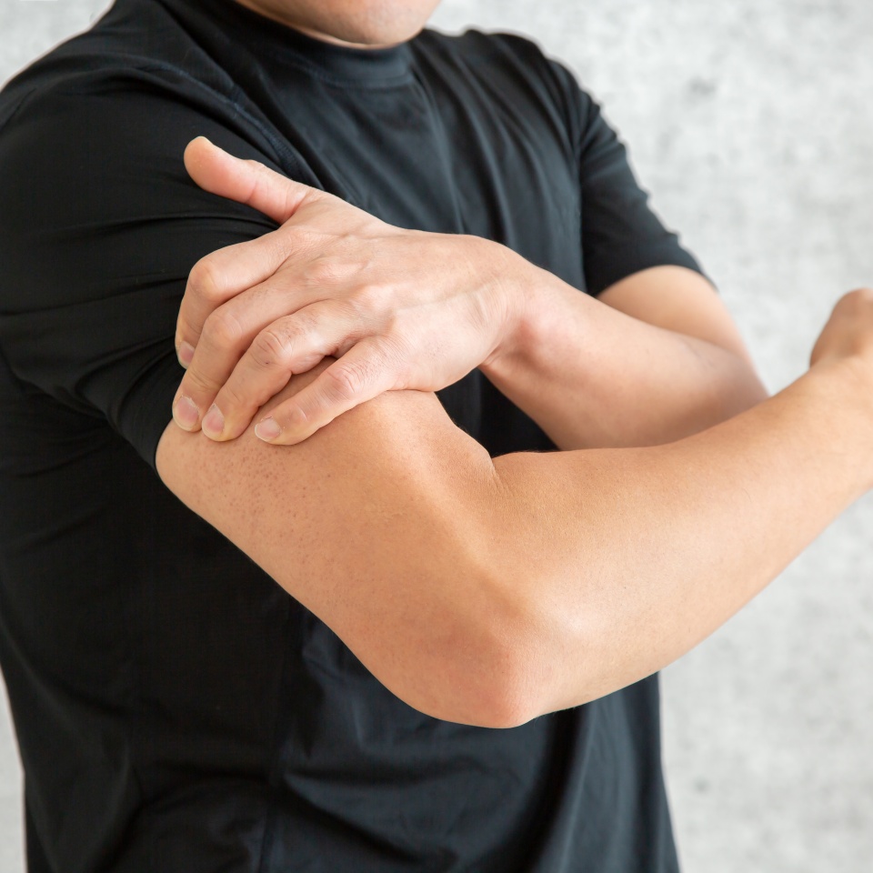 A person in a black t-shirt holds their left upper arm with their right hand, appearing to touch or massage the area, possibly due to discomfort or pain. The background is out of focus.