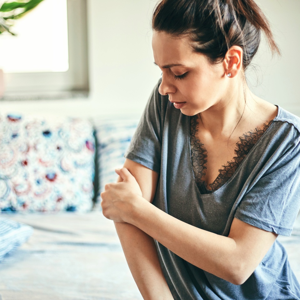 A woman with dark hair in a loose ponytail sits indoors, looking down while touching her right arm with her left hand. She is wearing a grey shirt with lace detailing at the neckline. A patterned sofa is in the background.