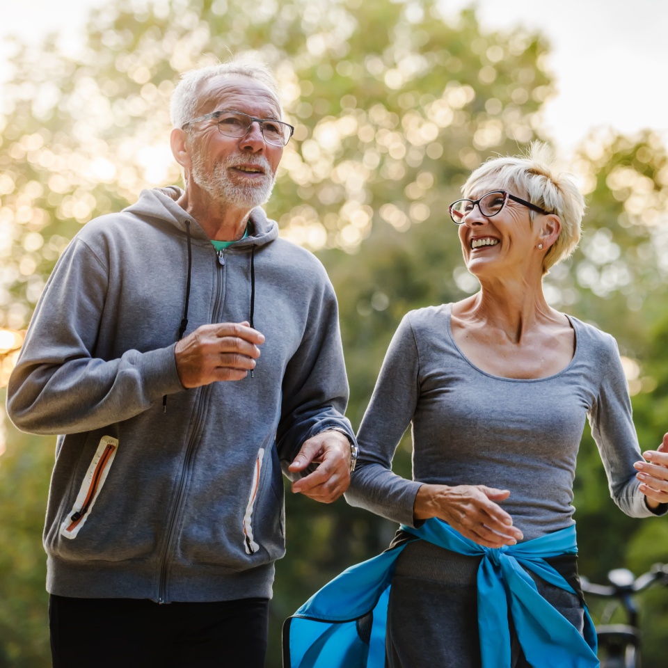 Two older adults wearing gray activewear are smiling and jogging together outdoors in a park, surrounded by greenery and warm sunlight.
