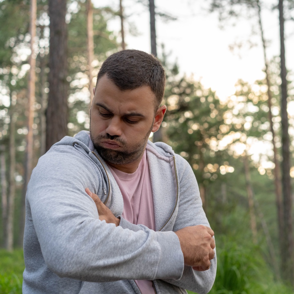 A man outdoors in a forest looks down at his upper arm with a displeased or pained expression while holding it with his opposite hand, as if he has discomfort or pain. He is wearing a gray hoodie over a pink shirt.