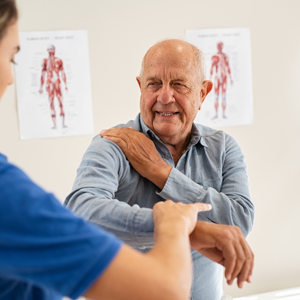 Young woman physiotherapist helping senior man with elbow exercise in clinic. Young woman doctor checking elbow of senior patient. Old man during an appointment with professional osteopath in private clinic working and massaging his shoulder pain.