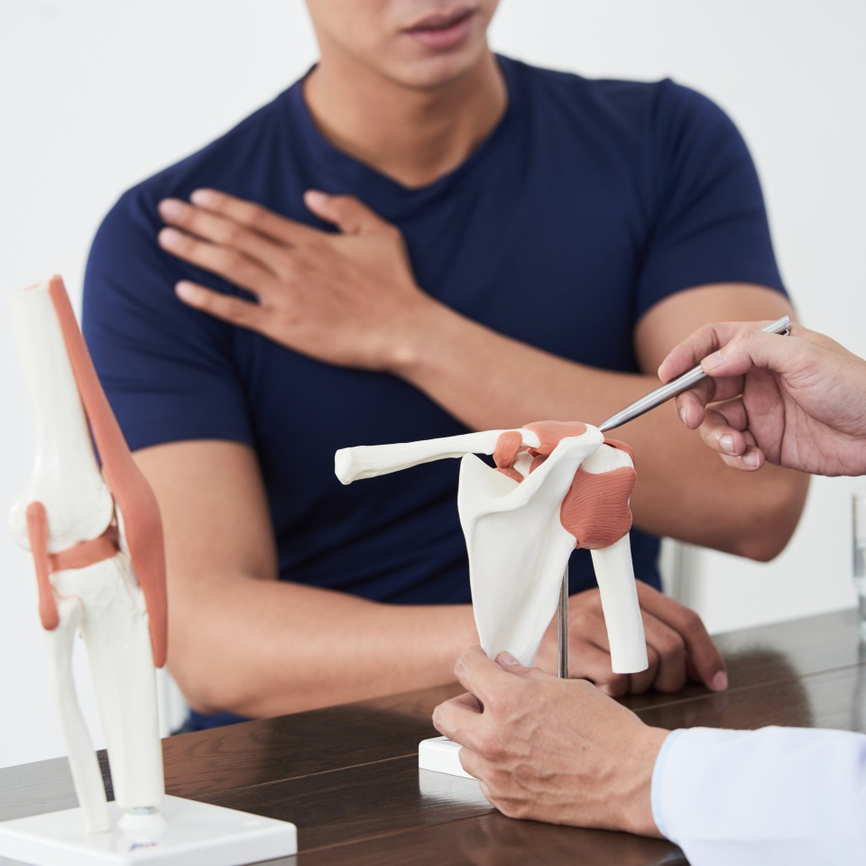 Horizontal shot of traumatologist sitting at desk in front of young Asian man consulting him about pain in shoulder joint