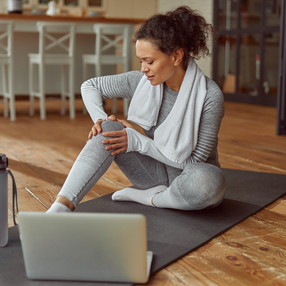 Female is sitting on mat and touching joint while suffering pain during video workout on laptop at home