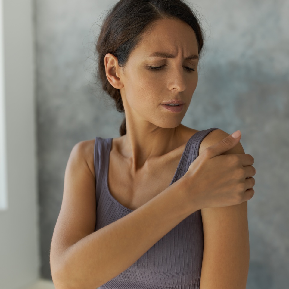 30-years-old good-looking dark-haired Caucasian woman in stylish violet fitness top standing in front of camera with painful face expression, looking at and stroking her shoulder over grey wall