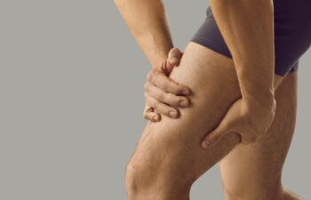 Man feeling pain or having thigh cramp. Sportsman who's suffering from hamstring muscles pain after running, jogging or gym fitness training standing on grey copy space background and touching his leg
