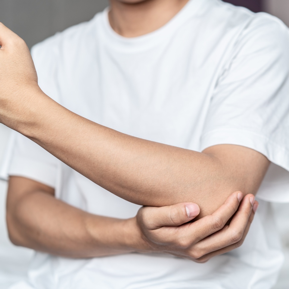 A person in a white shirt holds their left elbow with their right hand, appearing to experience discomfort or pain in their arm.