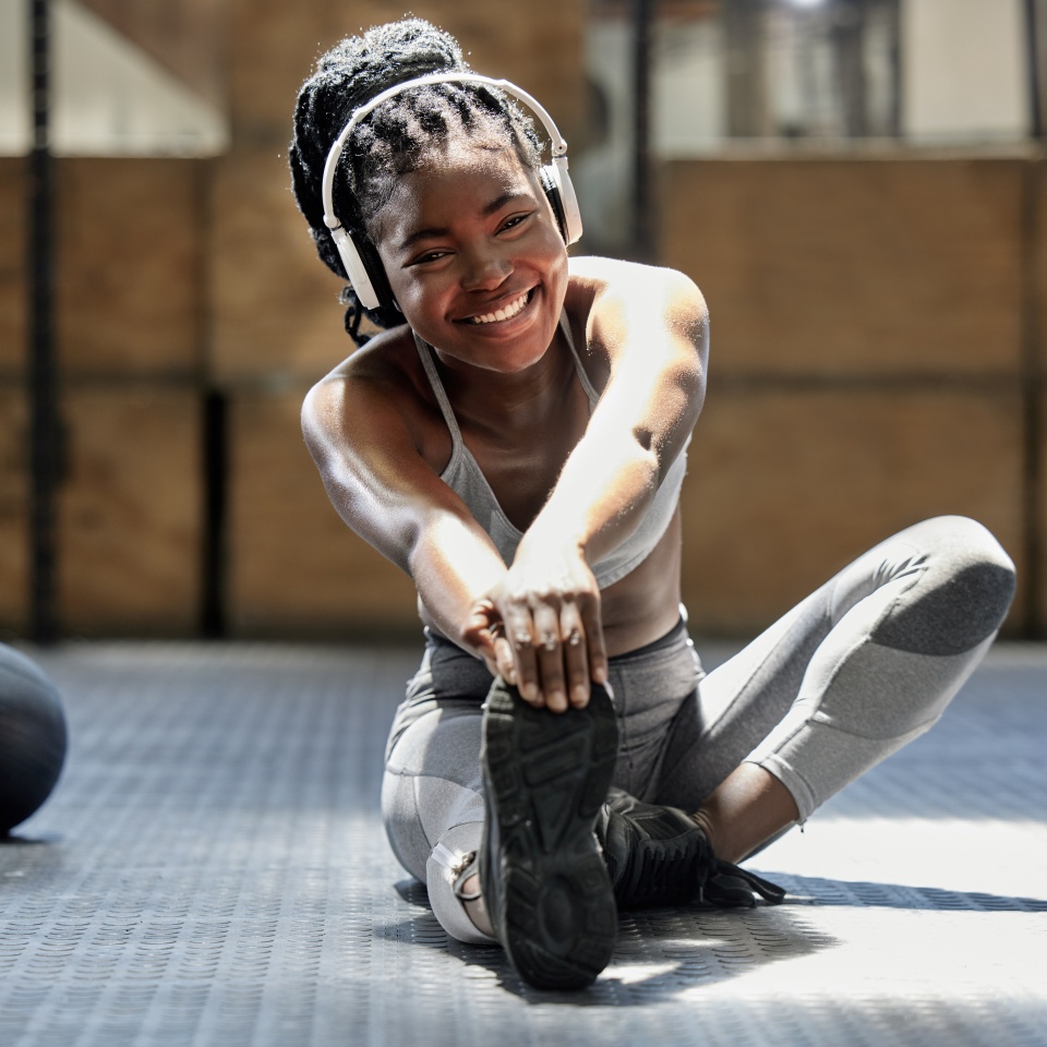 Portrait of woman stretching with music at gym, audio podcast for workout motivation and happy about fitness training on floor at health club. African athlete or sports person doing exercise.