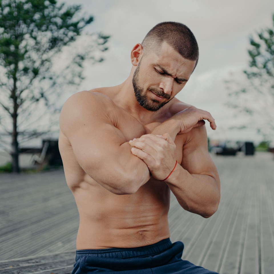 Outdoor shot of muscular guy injured elbow during physical exercises, feels strong pain and poses with naked torso. Health problems, medical, sickness and sport concept. Pain relief. Muscle injury