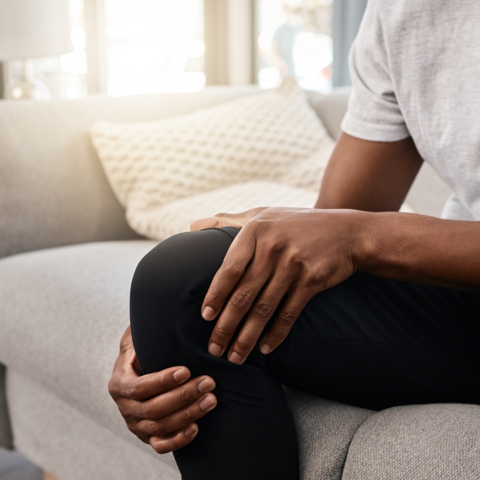 Knee pain, hand of a man and injury while sitting on home living room sofa for rest and relaxation. Ache, painful leg joint and african american man touching his knees for a strain muscle.