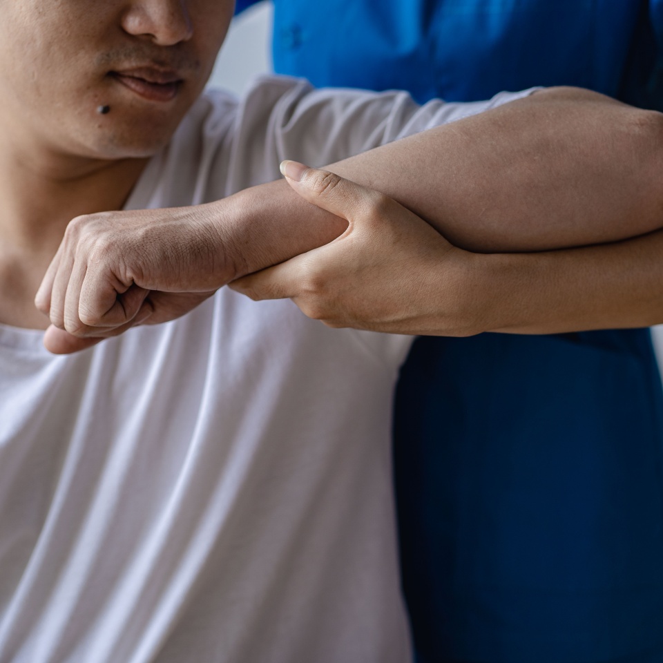 A person in a white shirt receives physical therapy, with another person in blue supporting and stretching their arm. The focus is on the hands and arm, highlighting the rehabilitation process.