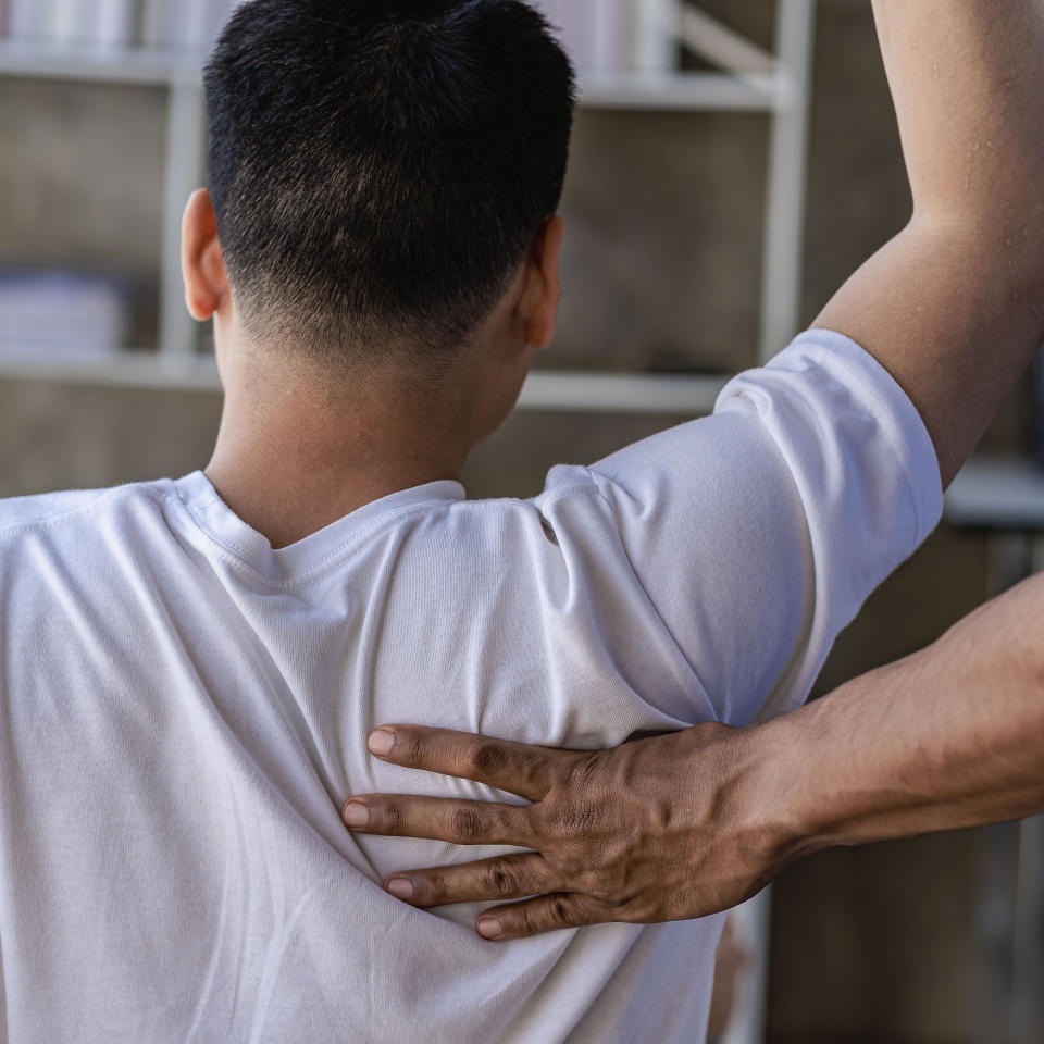 A person in a white shirt is receiving physical therapy, with a therapist’s hand supporting their upper back and shoulder. The person’s arm is raised as part of the therapeutic exercise.