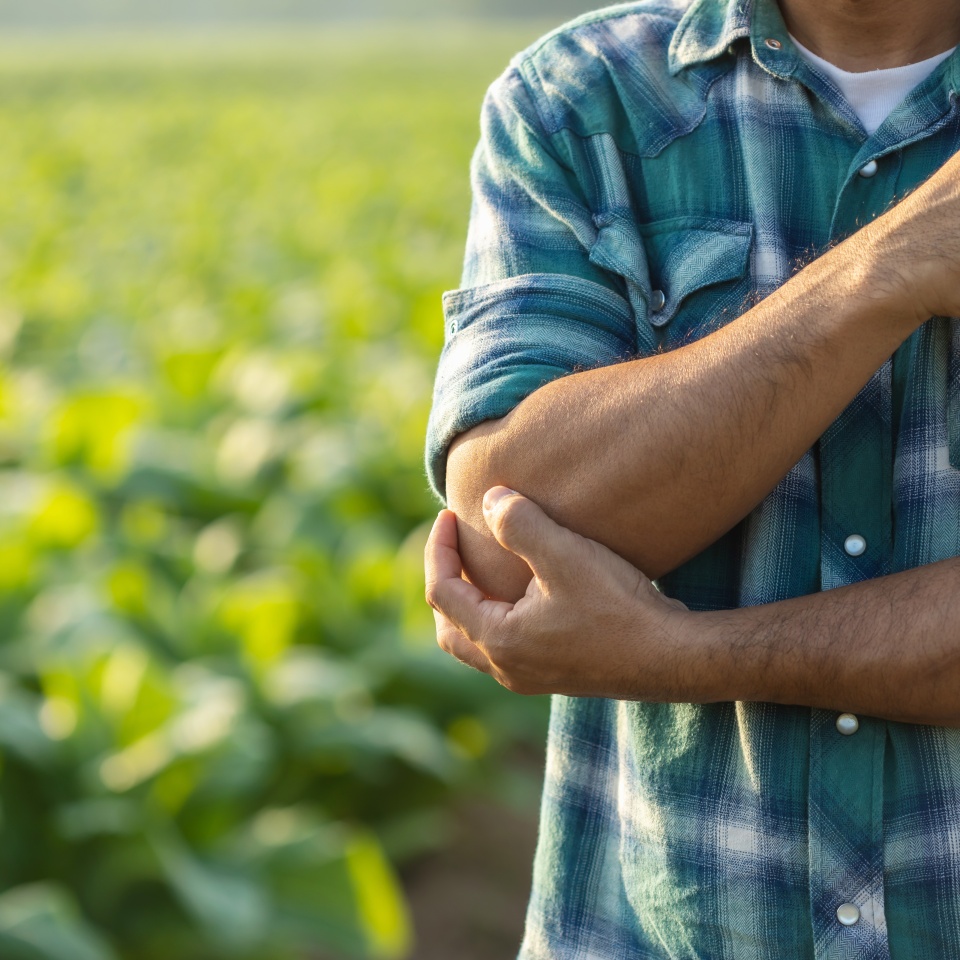 Injuries or Illnesses, that can happen to farmers while working. Man is using his hand to cover over elbow because of hurt, pain or feeling ill. Health of agriculturist concept.