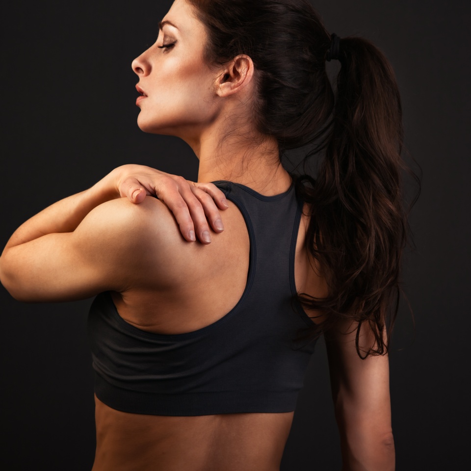 Female sporty muscular with ponytail doing stretching workout of the shoulders, blades in sport bra, holing the neck the hand on dark background with empty copy space. Back view. Healthy studio portrait