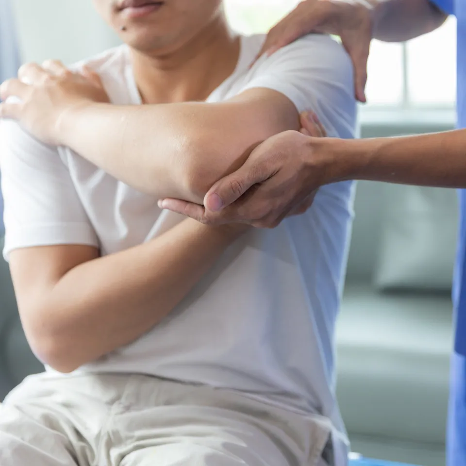 A person sits with arms crossed over their chest while another person in blue assists by supporting their elbow, possibly during a physical therapy or medical examination session.