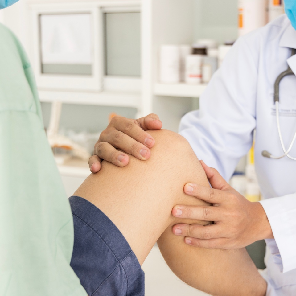 A doctor wearing a white coat and face mask examines a patients knee in a medical office. The patient is seated and wearing shorts, while the doctor gently holds and inspects the knee.