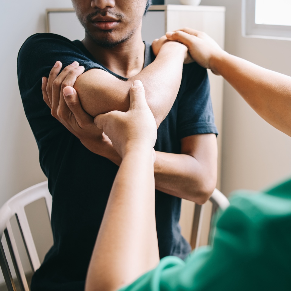 A person sits on a chair while another person stretches their arm across the chest, possibly performing a physical therapy or rehabilitation exercise.