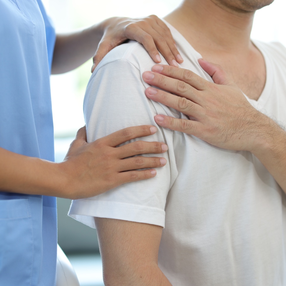 Doctor diagnosing man's shoulder pain in hospital examination room. Nurse doing massage and physiotherapy to treat patient's shoulder.