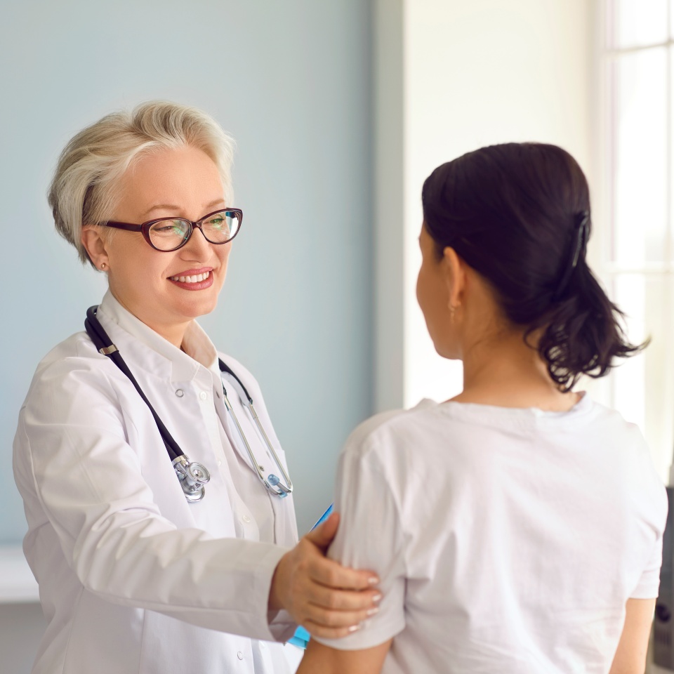 Doctor at the clinic or hospital supporting and reassuring her patient. Happy senior doctor in a white lab coat smiling and touching the young woman's shoulder. Medicine and healthcare concept