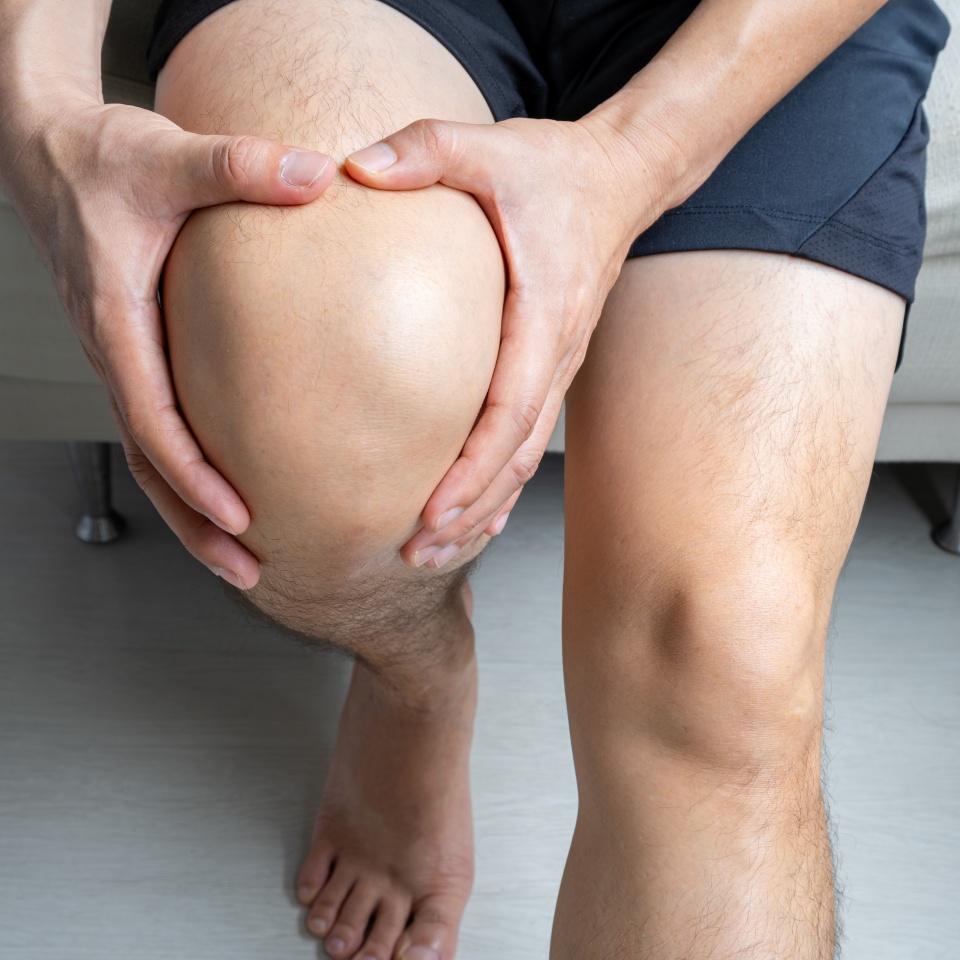 A person sitting down, wearing black shorts, is holding their left knee with both hands, suggesting pain or discomfort in the knee area.