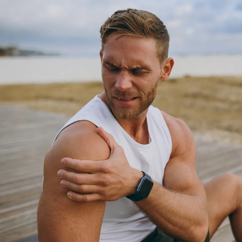 Young strong toned fit sportsman man wears sports clothes feels bad suffer from pain spasm hold shoulder warm up train at sunrise sun dawn over sea beach outdoor on pier seaside in summer day morning